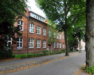 Blick auf die Volkshochschule, in der auch das Amt für Kinder, Jugend, Schule und Sport untergebracht ist. Es handelt sich um ein rot geklinkertes Gebäude mit zwei Reihen großer weißer Fenster und grünen Bäumen davor.