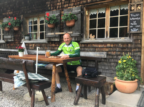 Thorsten Rodtgardt sitzt vor einer Bergalm an einem rustikalen Holztisch und hält ein Glas in der Hand. Er trägt Fahrradbekleidung.