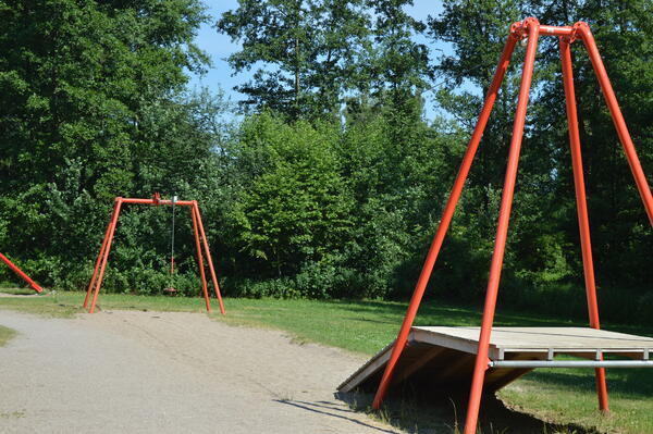 Spielplatz Zum Krückaupark. Auf einem Sandplatz an einer Wiesen neben Bäumen und Büschen ist eine Seilnahn mit roten Stangen.