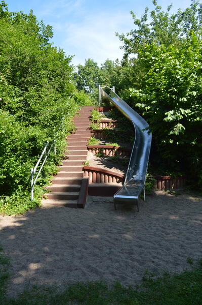 Eine Rutsche führt einen Hügel herunter. Nebenan ist iene Treppe neben ein paar Büschen.