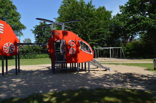 Ein Klettergerüst in Form eines Helikopters auf ienem Sandplatz. Im Hintergrund sind Schaukeln.
