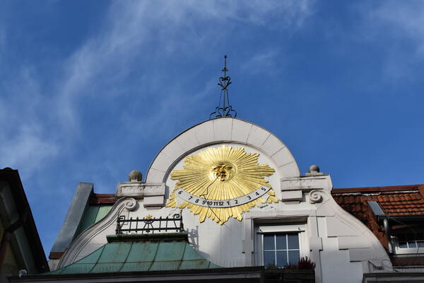 Golden prangt die Sonnenuhr an der Spitze der Jugendstilfassade, hervorgehoben vom blauen Himmel.