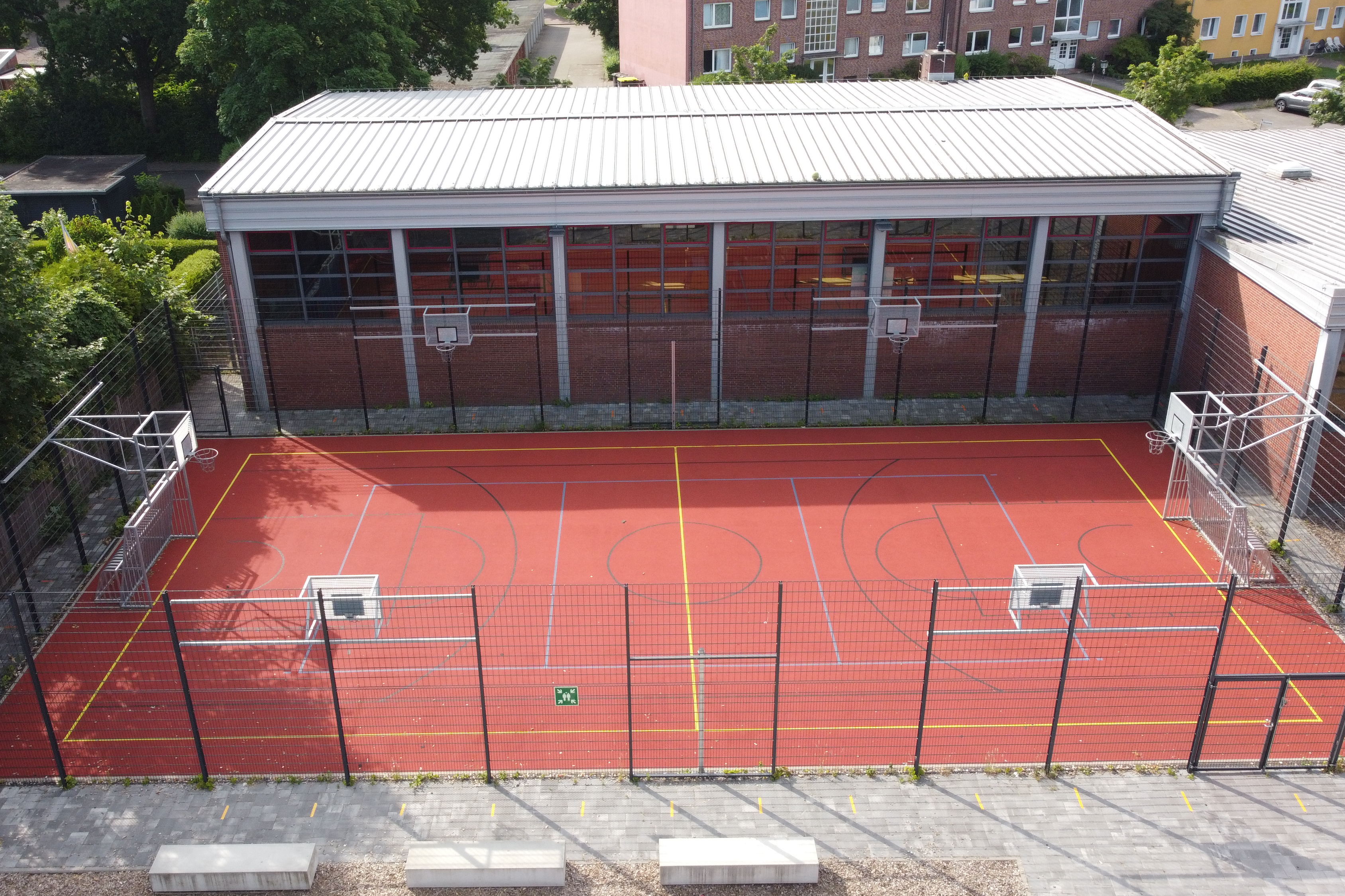 Auf einem eingezäunten Sportplatz stehen Basketballkörbe und Fussballtore. Nebenan ist die Sporthalle.