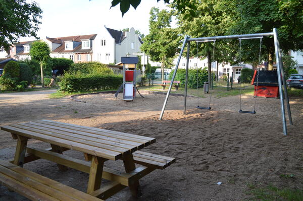 Neben einer Bank stehen auf dem Sandplatz joch ein kleines Gerüst mit Rutsche und zwei Schaukeln.