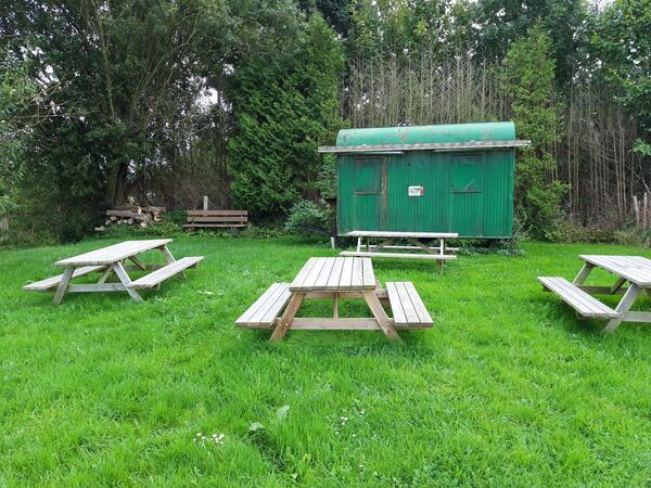 Bild vergr&ouml;&szlig;ern: Auf einer gr&uuml;nen Wiese stehen vor einem Bauwagen Parkb&auml;nke mit Tischen f&uuml;r Besucher bereit.