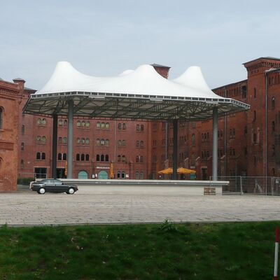 Ein gro&szlig;er, moderner Pavillion steht auf dem gepflasterten Vorplatz eines historischen Geb&auml;udes aus rotem Backstein.