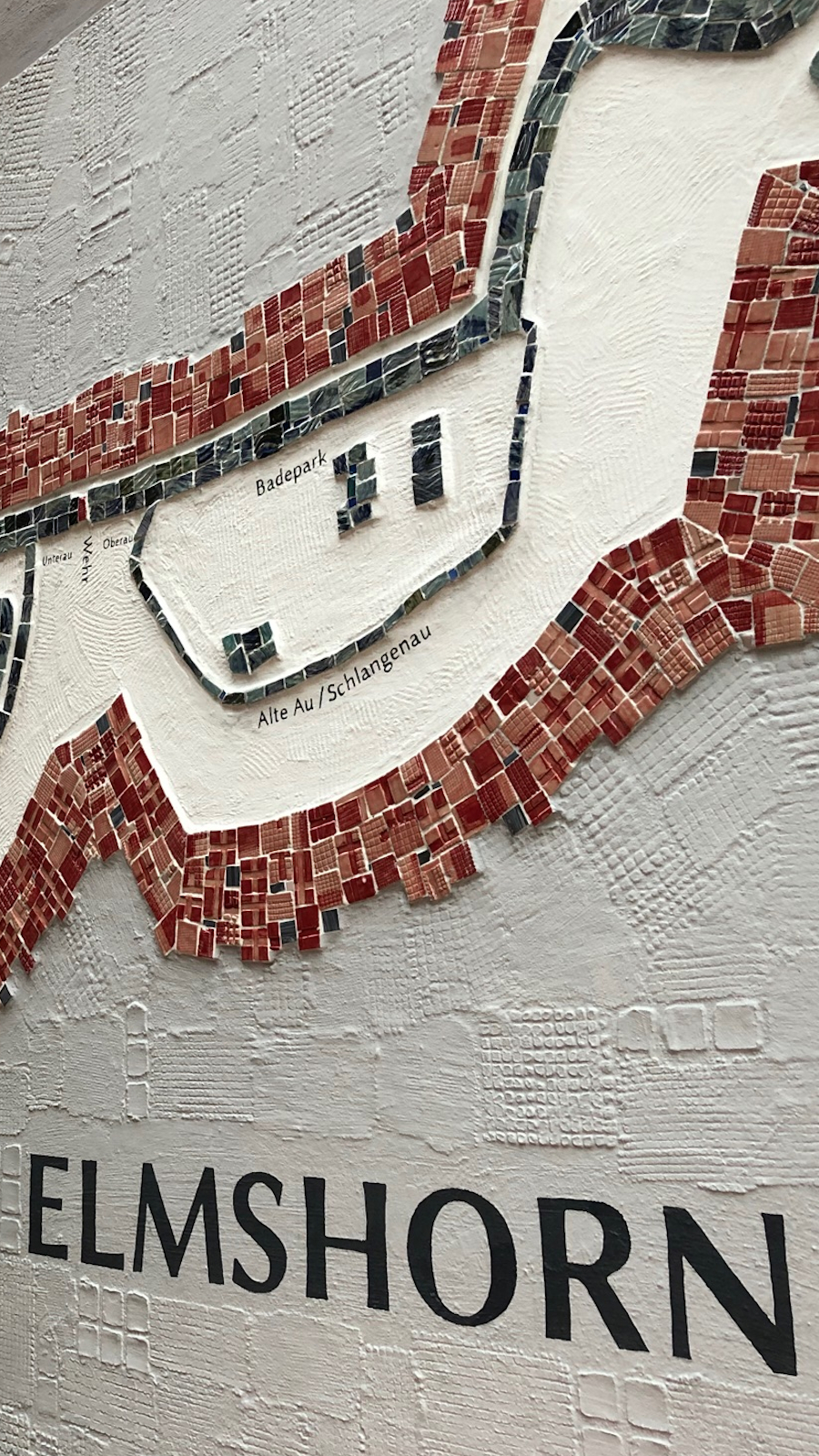 Das Kunstwerk der Künstlerin Ruth Alice Kosnick trägt die Bezeichnung Lauf der Krückau. Zu sehen ist eine Wandapplikation aus Mosaik mit Relief entlang einer Hauswand.