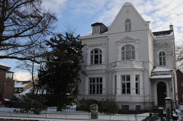 Vorderansicht einer weißen, mehrgeschossigen Villa hinter einem eingezäunten Vorgarten im Winter.