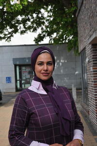 Soaad Ibrahim steht im Freien vor Gebäuden und lächelt für ein Porträtfoto in die Kamera. Sie trägt ein modisches blaues Kopftuch und einen weiß-karierten blauen Rock über einem hellen Hemd.