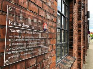 Bild vergr&ouml;&szlig;ern: Eine Glastafel mit einer Erl&auml;uterung in wei&szlig;er Schrift ist an der Backstein-Fassade des Industriemuseums angebracht.