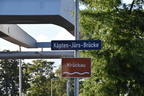 Die Käpten-Jürs-Brücke ist eine Klappbrücke die über die Krückau führt. Ein Schild mit dem Namen der Brücke und ein Schild mit dem Namen der Krückau befinden sich an einem Mast.