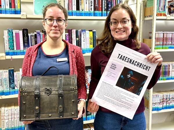 Die beiden Frauen stehen mit einer Schatzkiste und einer Zeitungsseite in der Hand vor Bücherregalen.